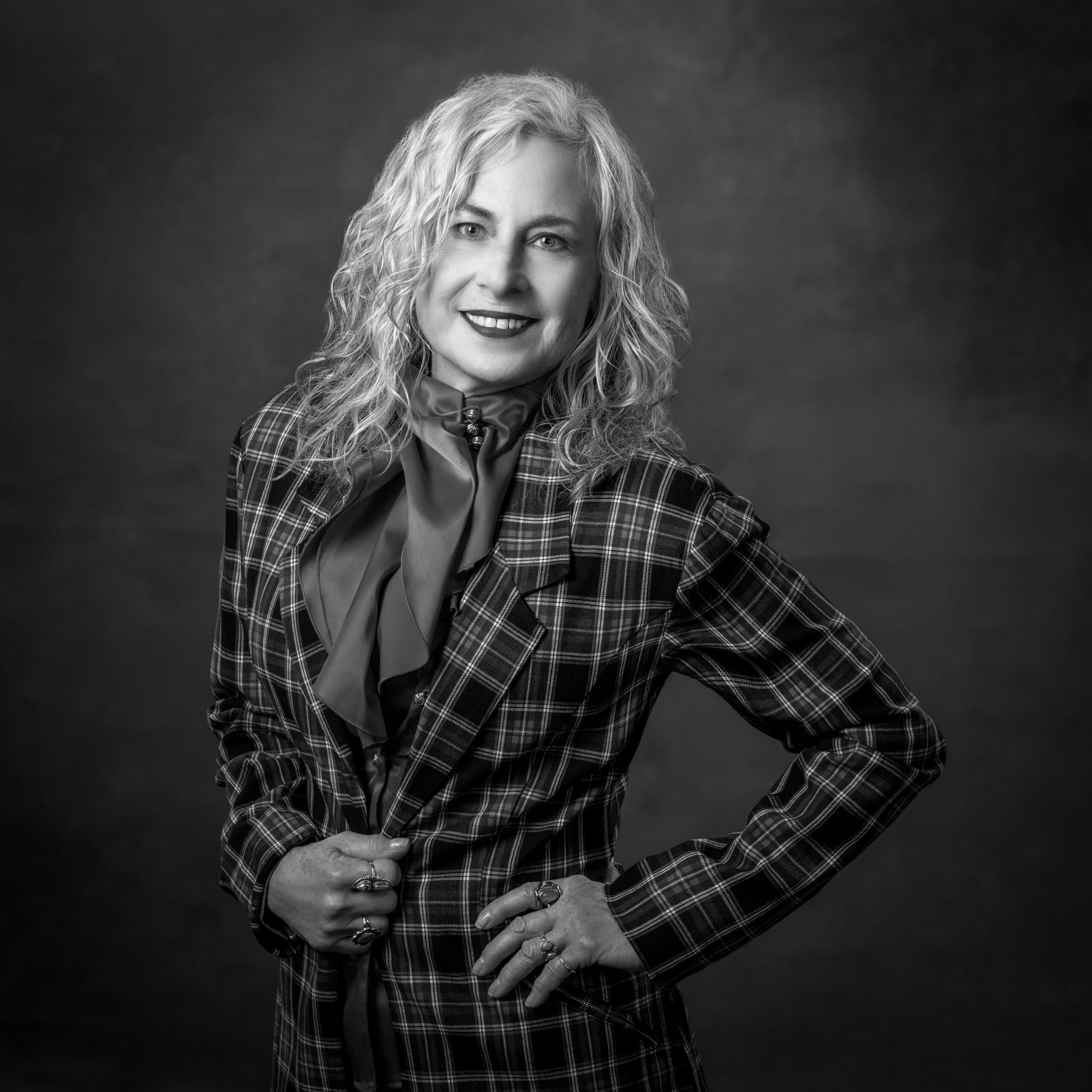 Woman with curly hair in plaid blazer, smiling, hands on hips, black and white portrait.