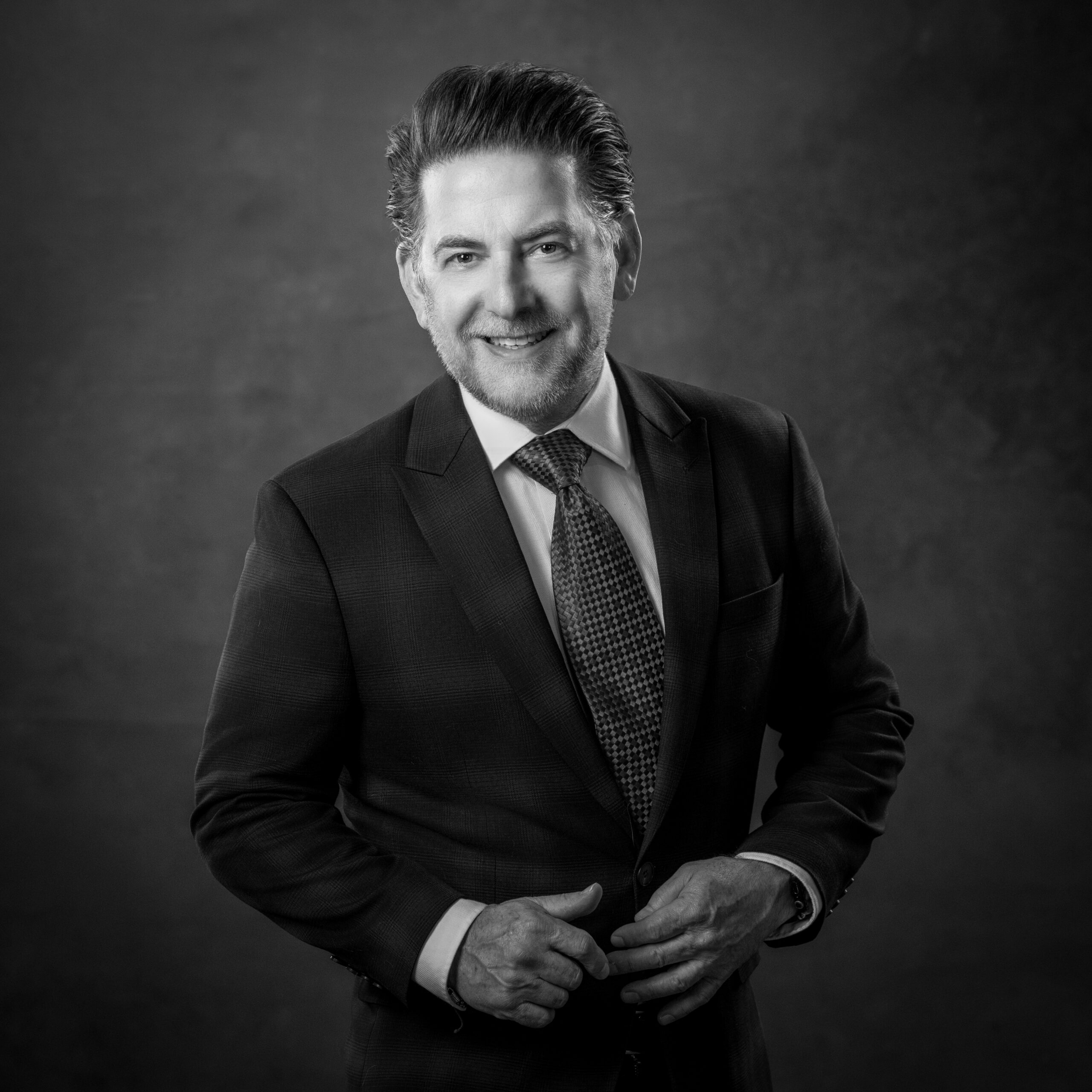 Smiling man in a suit and tie, buttoning his jacket, against a dark background.